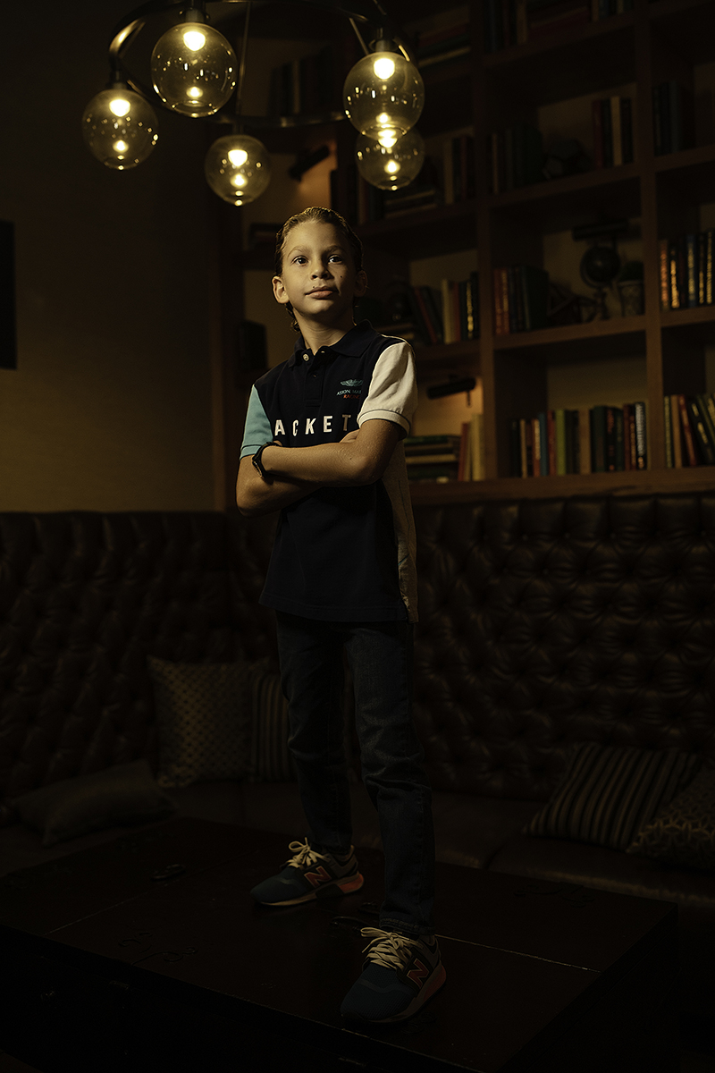 Menino em pé, confiante, sobre uma mesa em uma sala bem iluminada, com estantes de livros e luminárias modernas ao fundo.