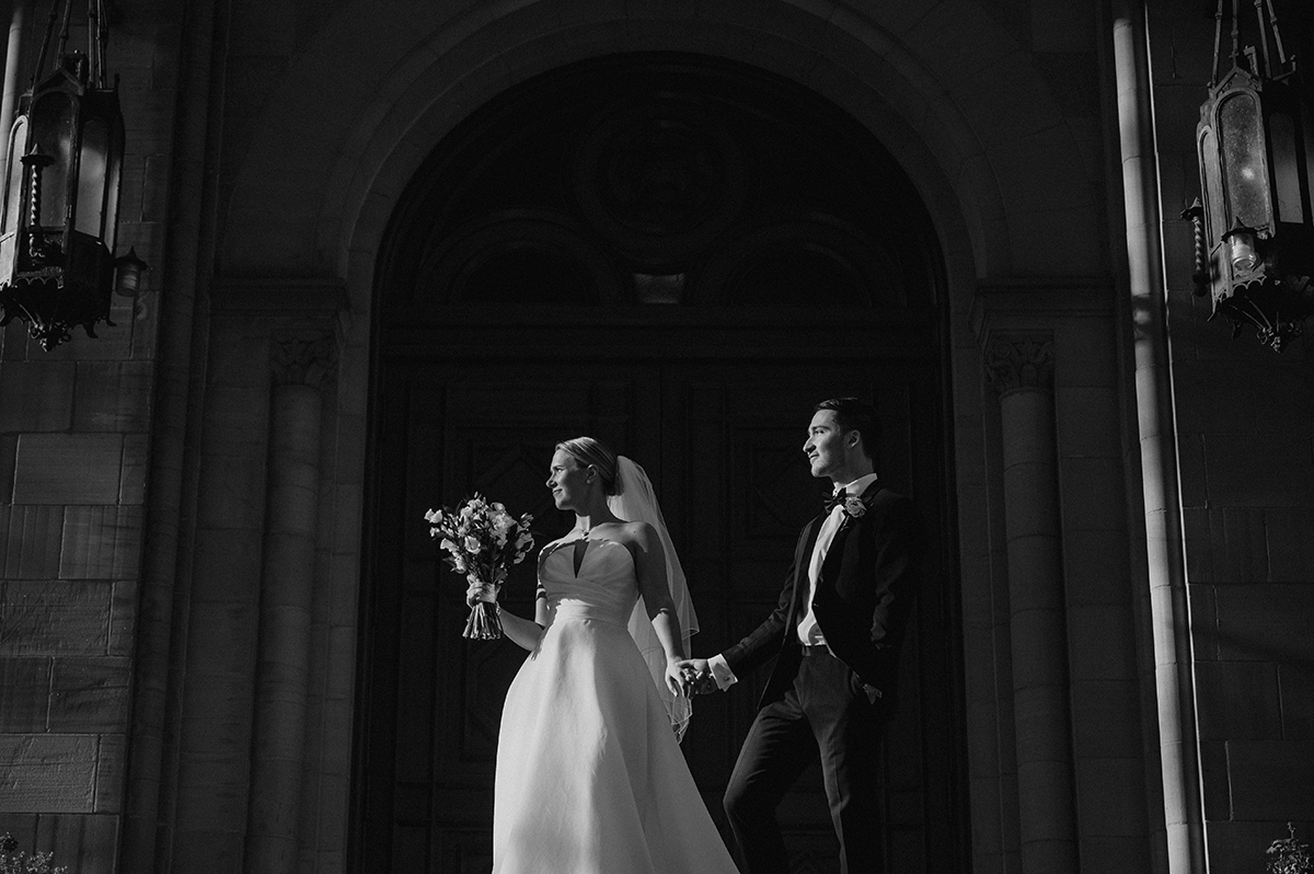 Les mariés se tiennent par la main à l'extérieur d'une cathédrale, magnifiquement éclairée en noir et blanc. La photo a été prise avec l'un des meilleurs objectifs de photographie événementielle pour les mariages. ©Rabih Madi