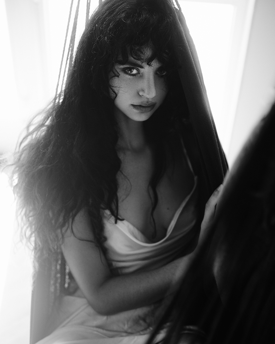 Femme aux longs cheveux bouclés, assise dans la lumière douce d'une fenêtre, regardant intensément l'appareil photo. Cette image en noir et blanc illustre les techniques d'éclairage de portrait pour le caractère, en mettant l'accent sur l'ambiance, la profondeur et le jeu d'ombres dramatique dans la photographie d'art.