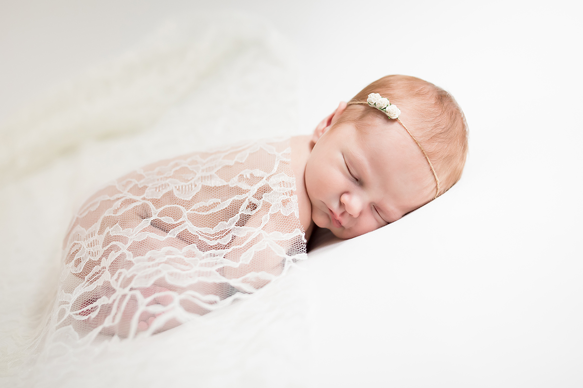 Bebê recém-nascido dormindo tranquilamente, enrolado em um cobertor de renda em um fundo branco - exemplo ideal de fotografia de retrato infantil com iluminação suave e profundidade de campo rasa.