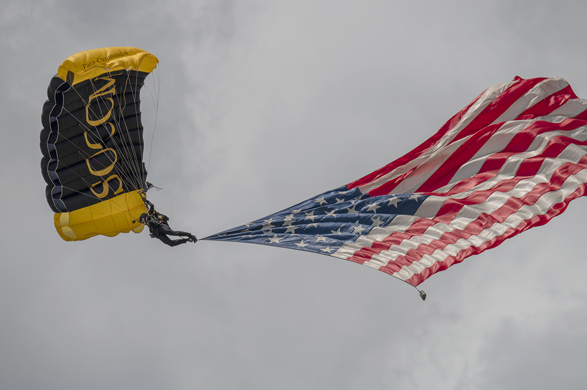 A SOCOM Para-Commando parachutist descends with a large American flag billowing beneath a yellow and black canopy against a cloudy sky.