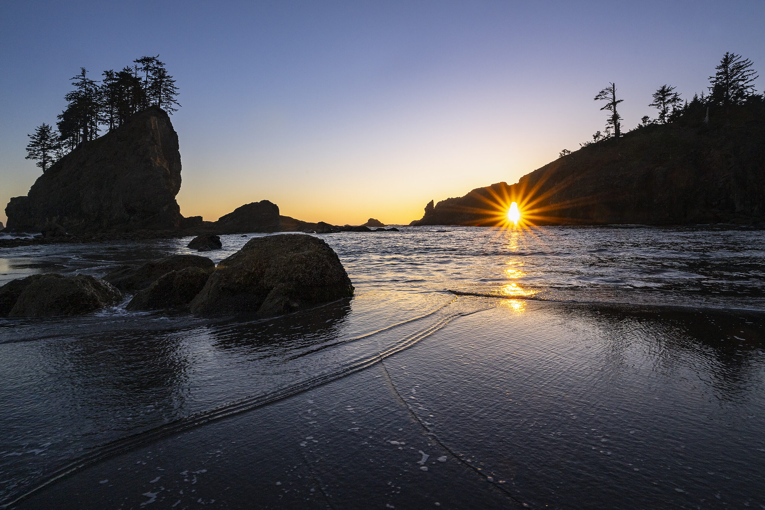 A coastal sunset with the sun perfectly aligned through a rock arch, casting a golden reflection on the ocean waves. Silhouetted sea stacks and trees frame the scene. Using a lens hood helps reduce glare and enhance contrast in this high-backlight composition.