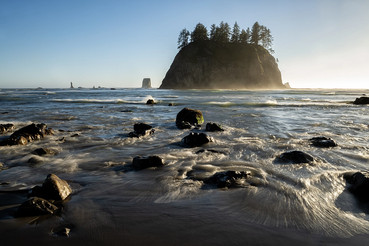 A coastal scene featuring an island with evergreen trees, surrounded by misty ocean waves and scattered shoreline rocks. The low-angle sunlight creates a dramatic effect, where using a lens hood helps reduce glare and maintain contrast in the highlights and shadows.