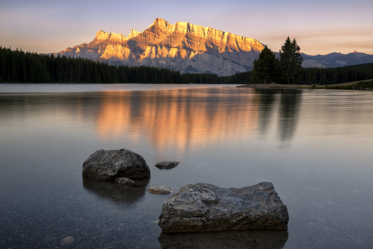 A peaceful lake with still water reflecting the golden glow of the sun on mountains, refined through long exposure photo editing.
