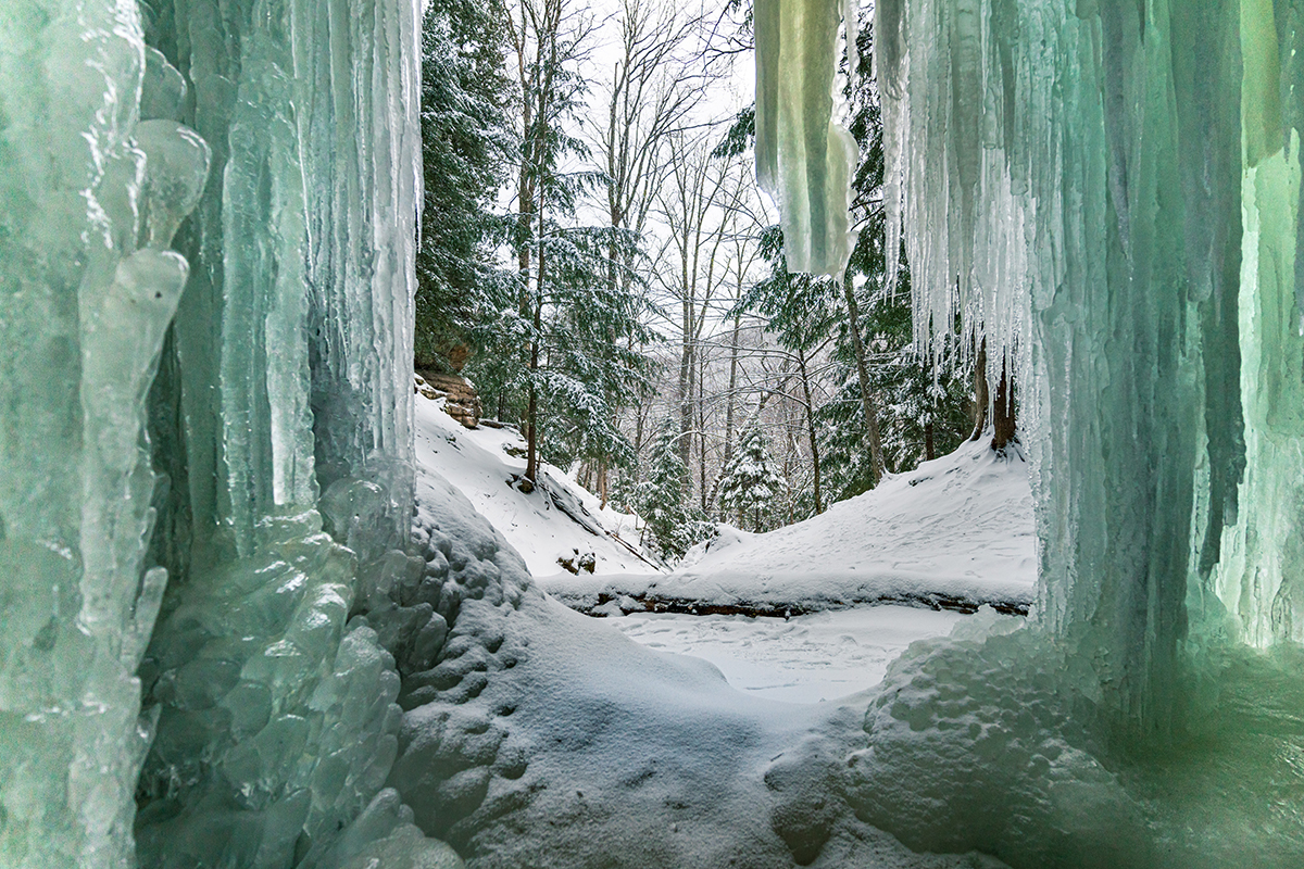 Vista através de enormes pingentes de gelo congelados que emolduram uma trilha de floresta nevada alinhada com sempre-vivas e árvores nuas de inverno.