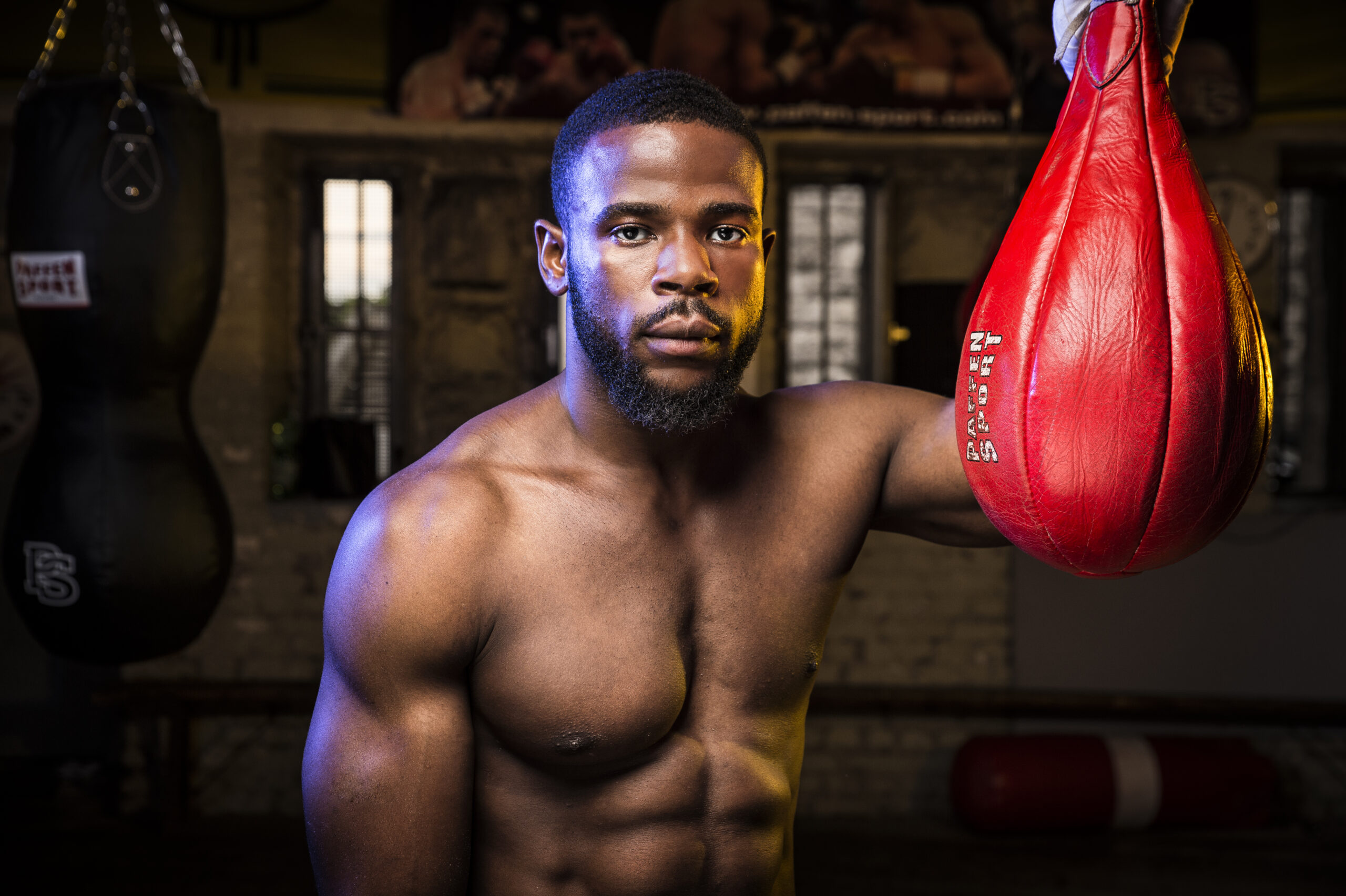 Un boxeador seguro de sí mismo posa sin camiseta junto a un saco rojo de velocidad en un gimnasio poco iluminado, con una iluminación espectacular que esculpe su físico y pone de relieve las técnicas expertas de fotografía de retratos con poca luz.
