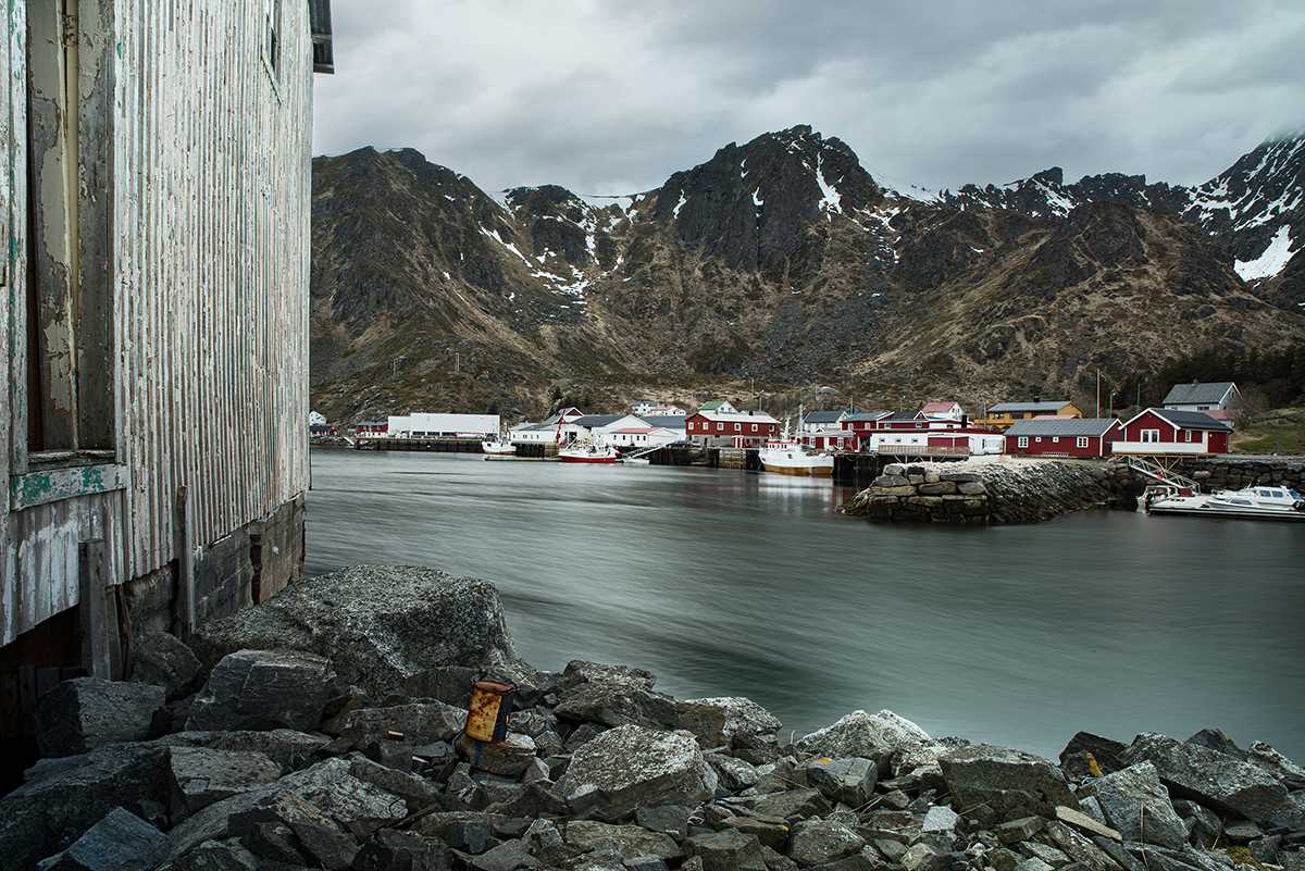 A small fishing village with red and white buildings dotted around a rocky shoreline with a wooden building in the foreground. 