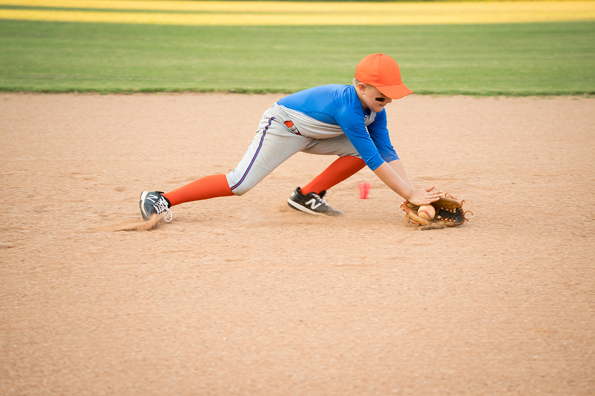 Un jeune garçon en uniforme de baseball s'étire pour attraper une balle au sol pendant un match, capturant parfaitement l'énergie et le mouvement des enfants en train de jouer - un exemple idéal de photographie d'enfants en mouvement.
