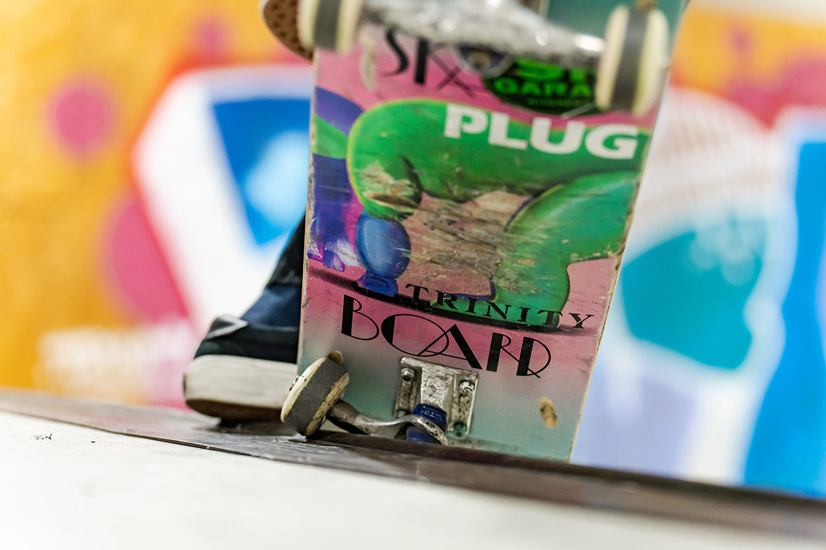 Primer plano de la colorida cubierta y los desgastados trucks de un monopatín encaramado al borde de una rampa, mostrando signos de uso frecuente.