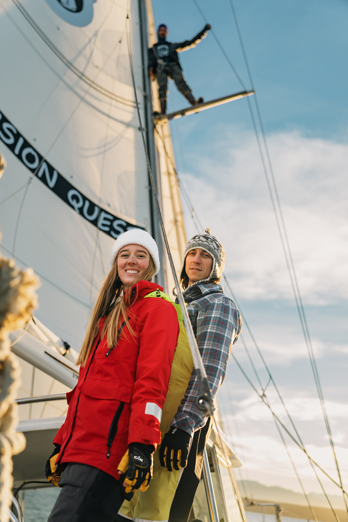 A group of sailors on a boat captured in candid style photography, with two smiling individuals in the foreground wearing winter gear and a third person high on the mast, set against a clear sky.
