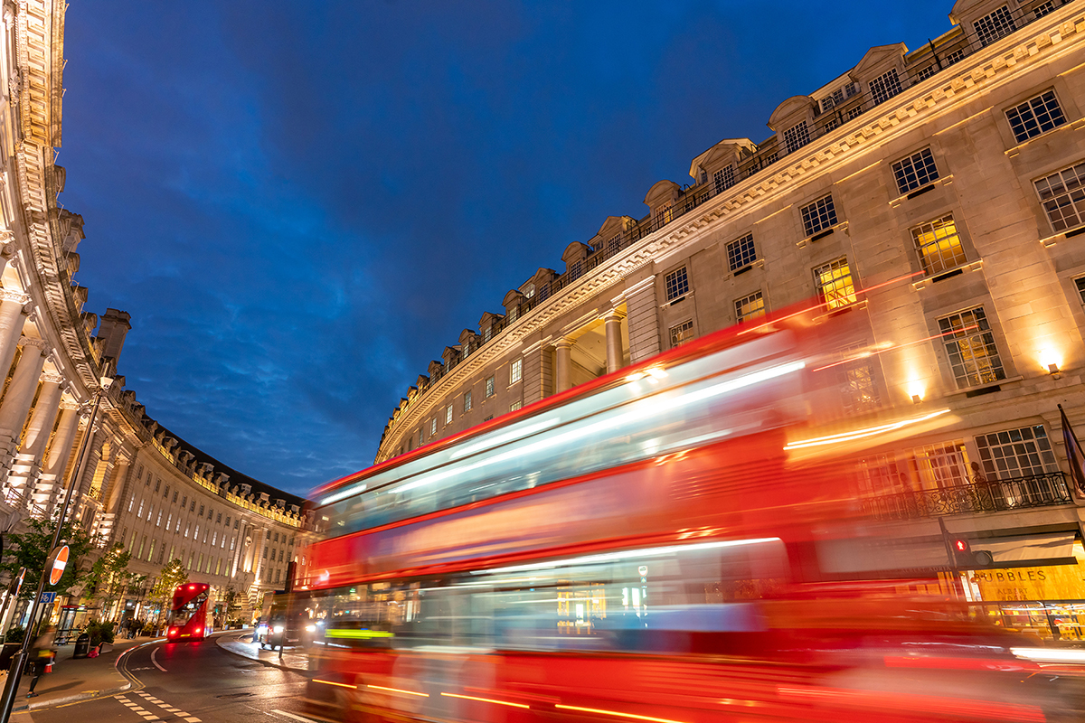 A red double decker bus passes through a vibrant and historic city street at night, shot using long exposure photography to enhance the motion blur effect.