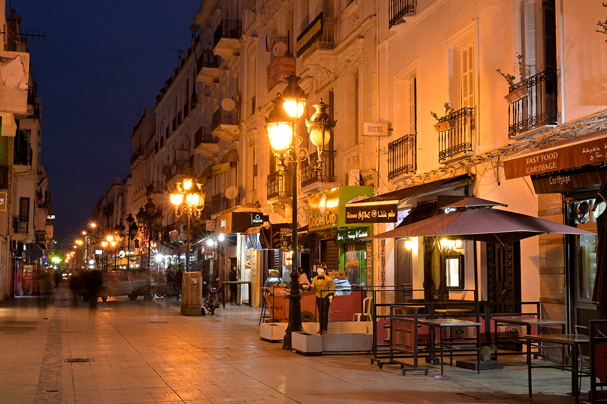 A shot of a quiet city street at night with sidewalk cafes and shops with colorful awnings illuminated by warm streetlights.