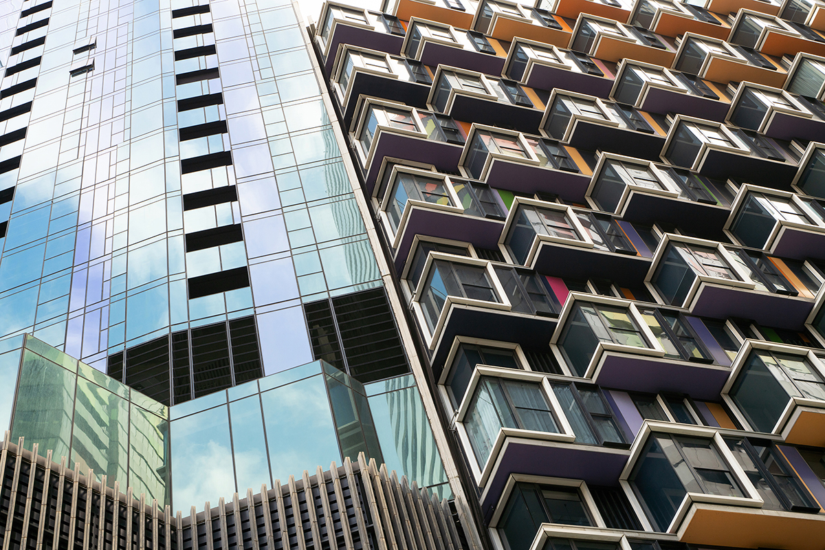 Uma foto dinâmica de paisagem urbana mostrando dois edifícios modernos contrastantes - um com elegantes painéis de vidro refletivo e outro com varandas arrojadas, geométricas e multicoloridas em forma de caixa - capturada de um ângulo baixo olhando para o céu.