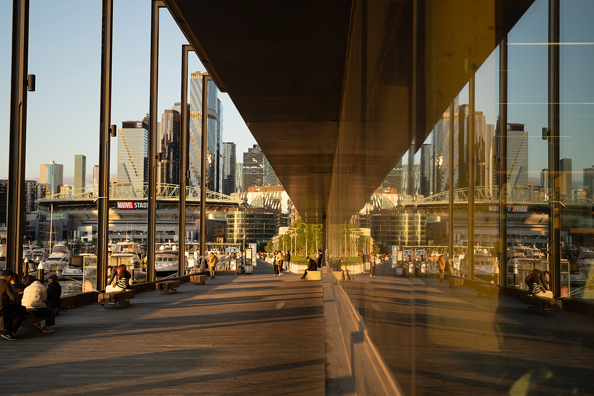 Fotografia da paisagem urbana de uma orla moderna durante a hora dourada, capturando um reflexo dramático dos arranha-céus e do Estádio Marvel na fachada de vidro de uma estrutura de calçadão.