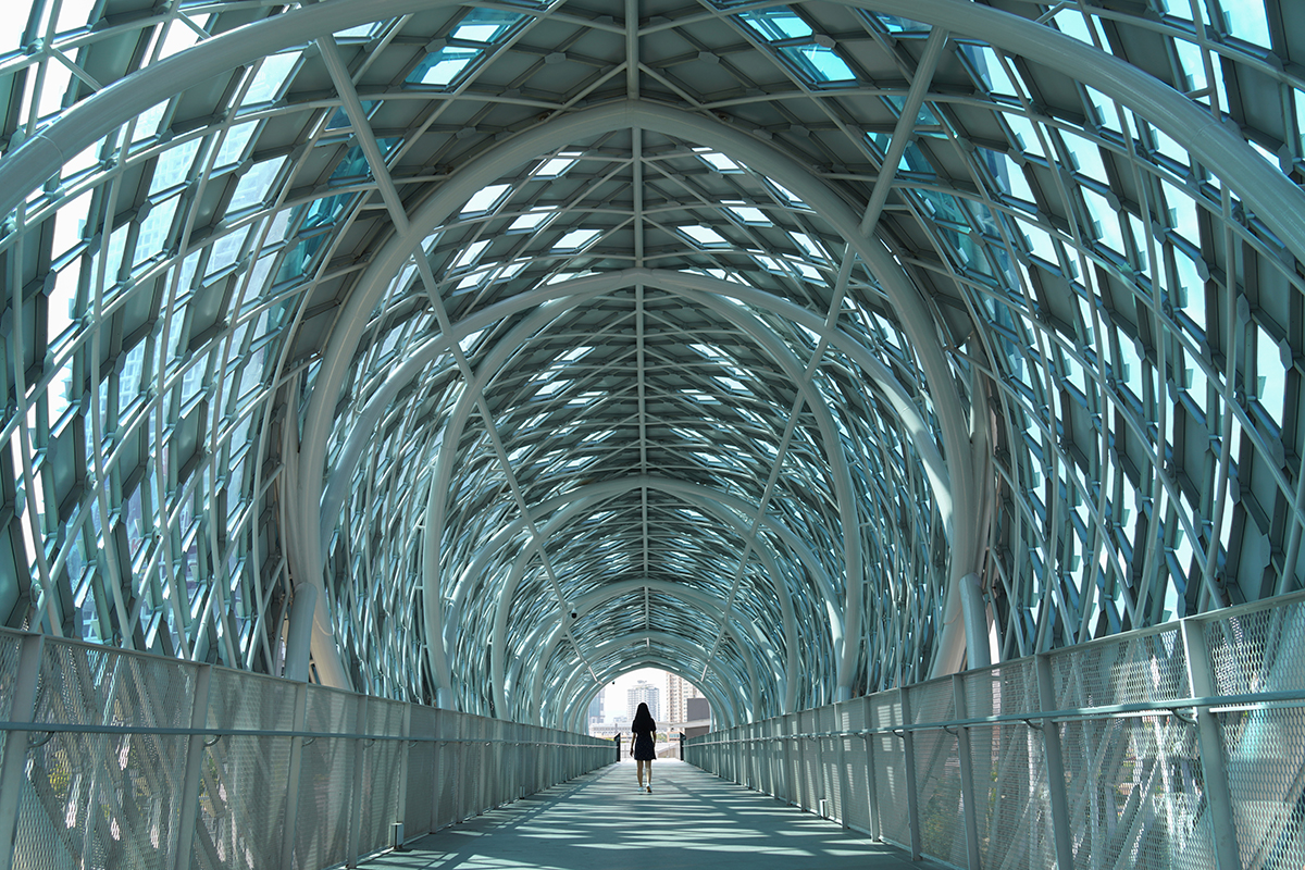 Túnel arquitetônico com fortes linhas de metal e vidro convergindo em direção a uma figura distante, ilustrando o uso de linhas de direção na fotografia para criar profundidade e guiar o olhar do observador.