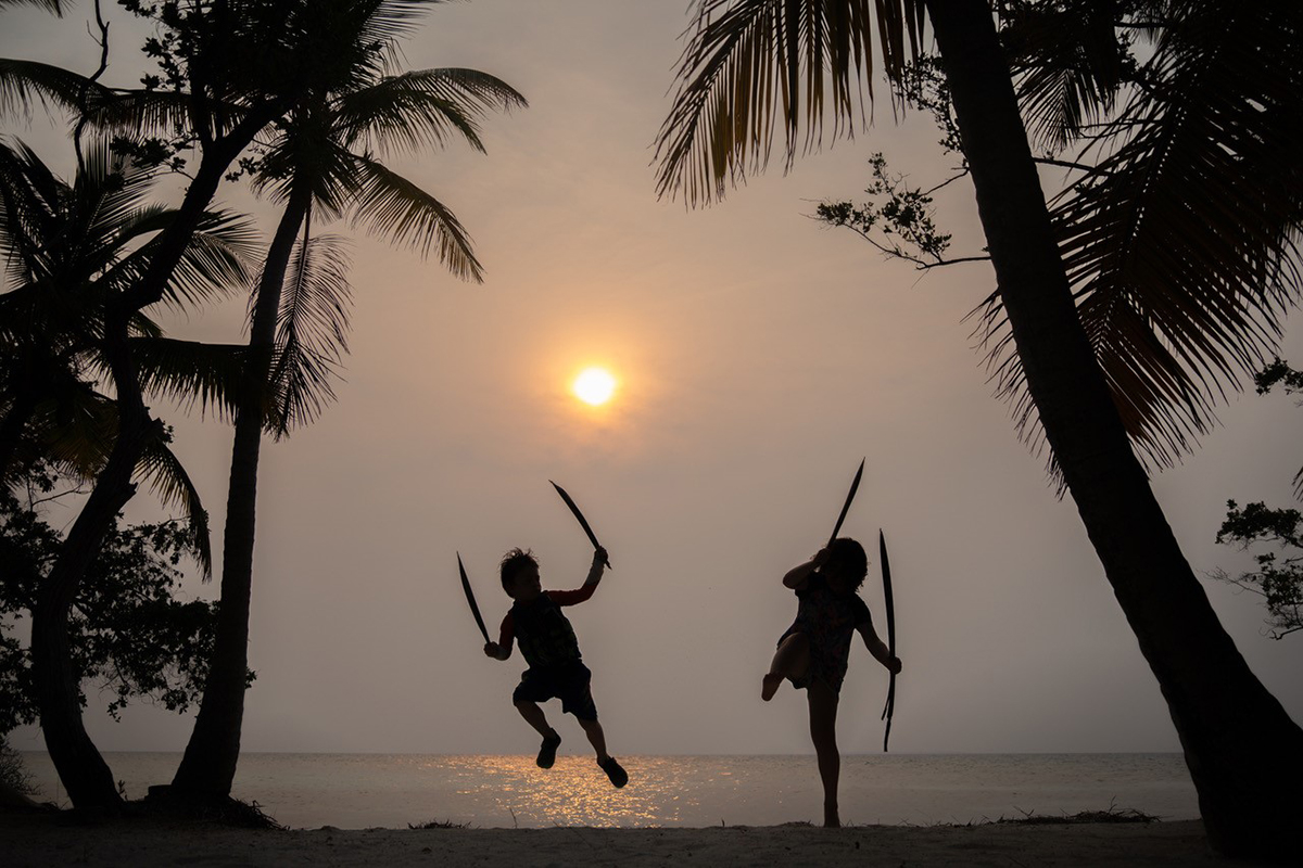 Siluetas de dos niños saltando con palmeras al atardecer en una playa tropical, capturando la acción lúdica y la atmósfera para mostrar cómo contar una historia con la fotografía. ©Andre Costantini