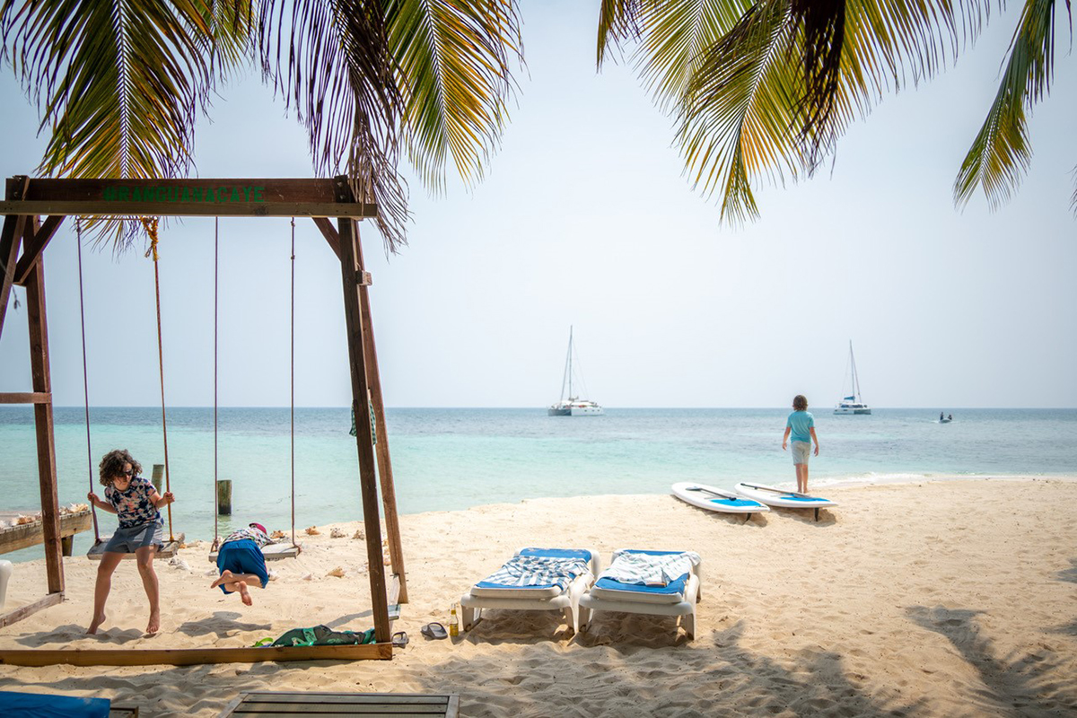 Niños jugando en un columpio de playa bajo palmeras mientras otro niño camina hacia la orilla con veleros a lo lejos en un día soleado. ©Andre Costantini