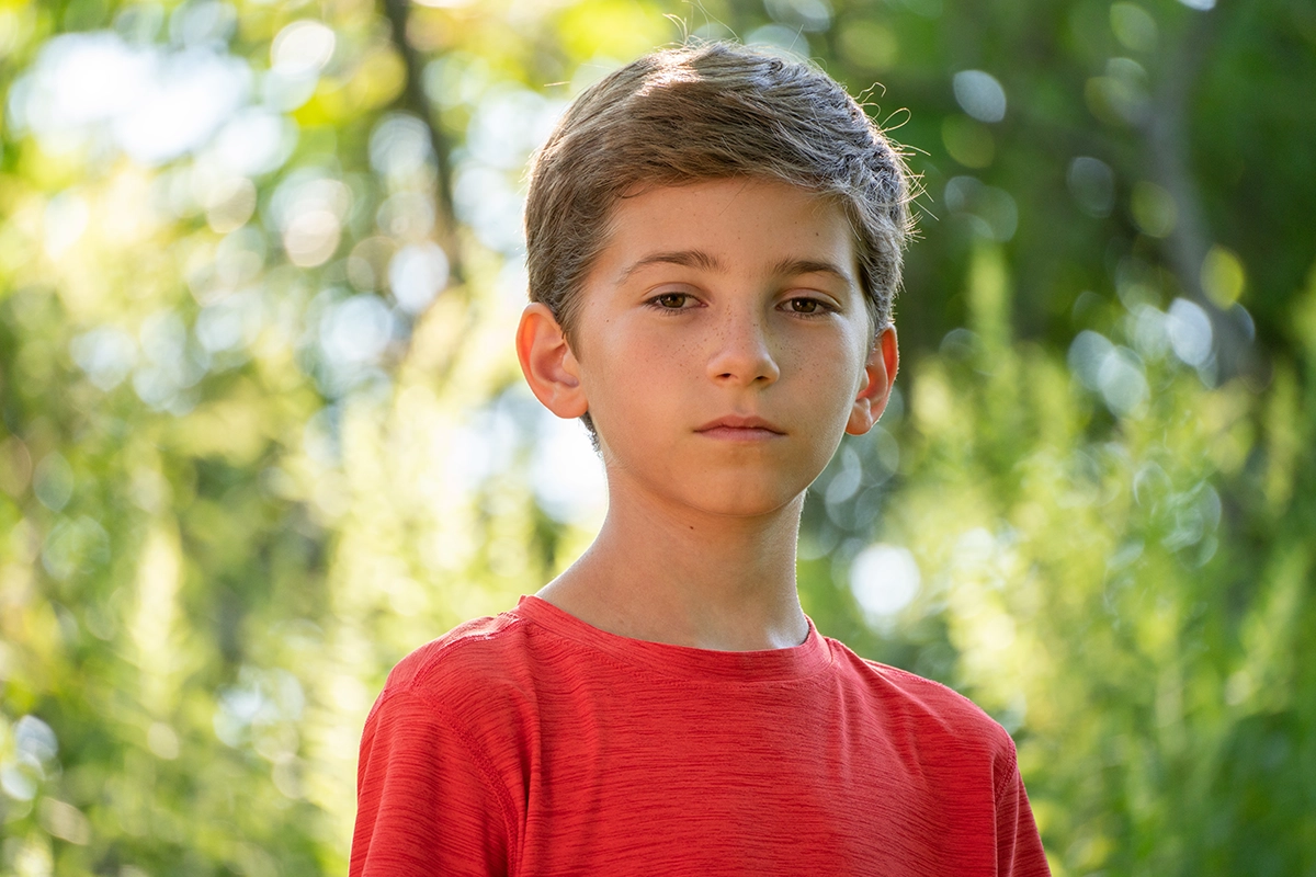 A portrait of a young boy in a red shirt, standing outdoors with a softly blurred green background. The sunlight filtering through the trees creates a natural bokeh effect. Using a lens hood helps control backlight and reduce lens flare, ensuring balanced contrast and clarity in the image.