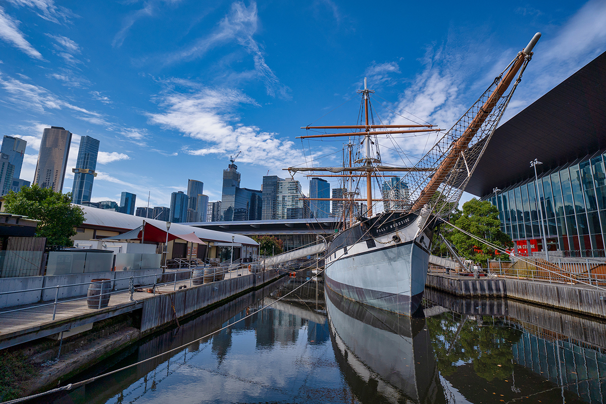 Fotografia de paisagem urbana capturando um contraste impressionante entre o histórico navio Polly Woodside e o pano de fundo dos modernos arranha-céus de Melbourne sob um céu azul vívido.