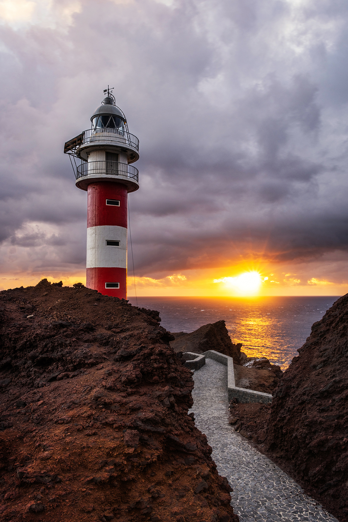 Caminho de pedra que leva a um farol vermelho e branco ao pôr do sol, ilustrando as linhas de direção na fotografia, guiando o olhar do observador para o ponto focal no horizonte.
