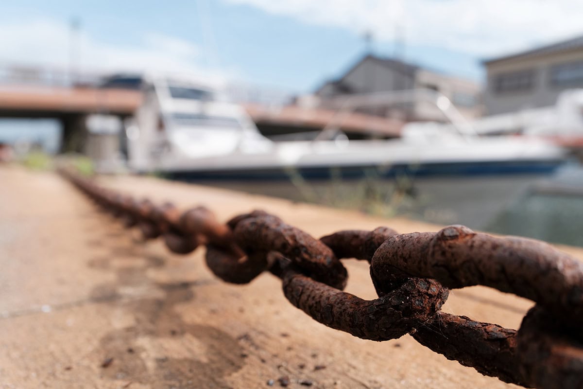 Primer plano de una cadena oxidada que se extiende a lo largo de un muelle con barcos y edificios suavemente difuminados al fondo en un día soleado.