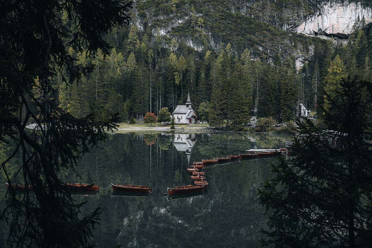 Lago alpino tranquilo com barcos de madeira e uma pequena capela refletida na água parada, emoldurada por árvores da floresta nas Dolomitas.