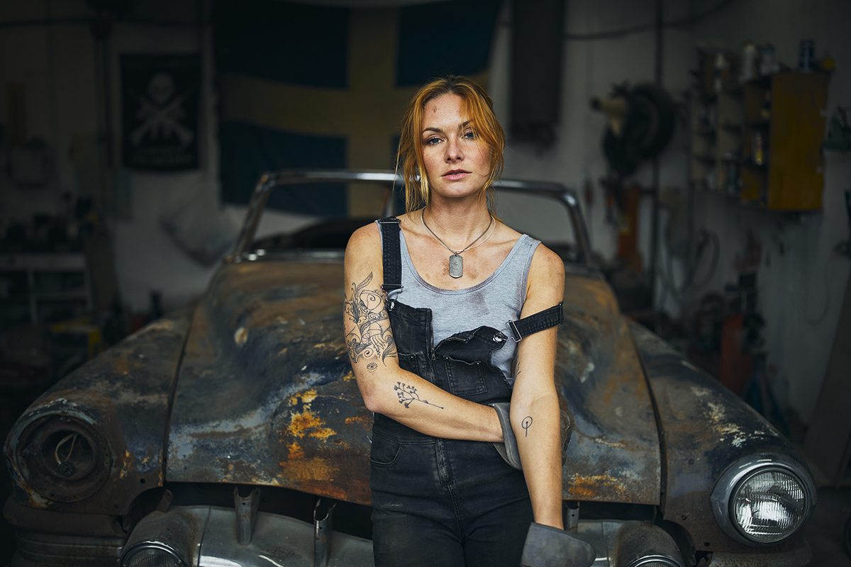 A gritty female mechanic stands confidently in front of a rusted vintage car inside a dimly lit garage&mdash;showing how to get a cinematic look through moody lighting, textured environments, and authentic character portrayal.