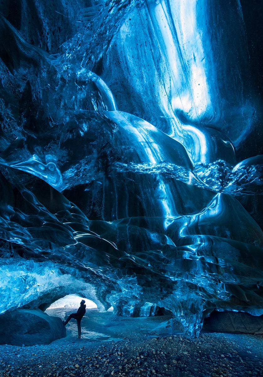 Silhueta de um caminhante dentro de uma caverna de gelo azul brilhante, ilustrando técnicas criativas de fotografia de inverno usando luz, escala e composição.