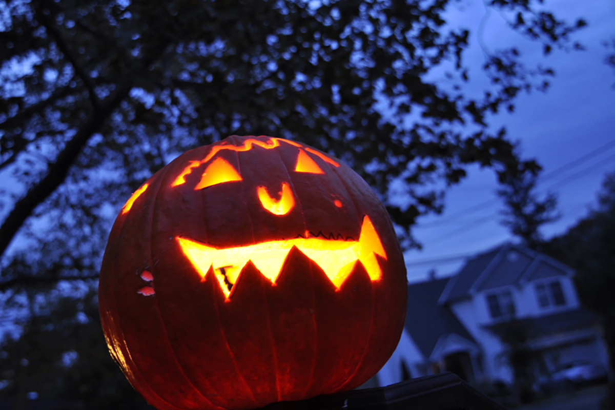 Photographing jack o&rsquo;lanterns at dusk &mdash; toothy pumpkin face glowing against a moody blue sky with suburban home in the background.