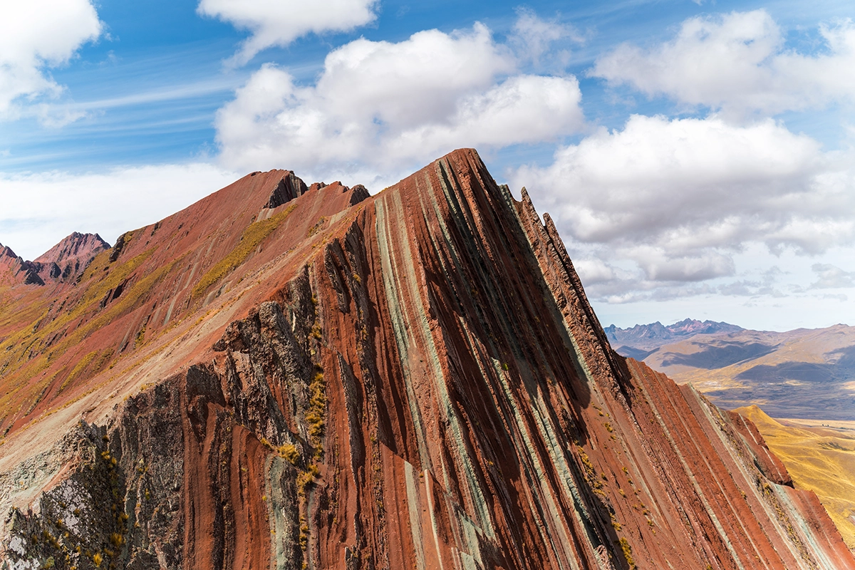 A striking mountain ridge with vibrant red, brown, and green striations under a bright blue sky with scattered clouds. 