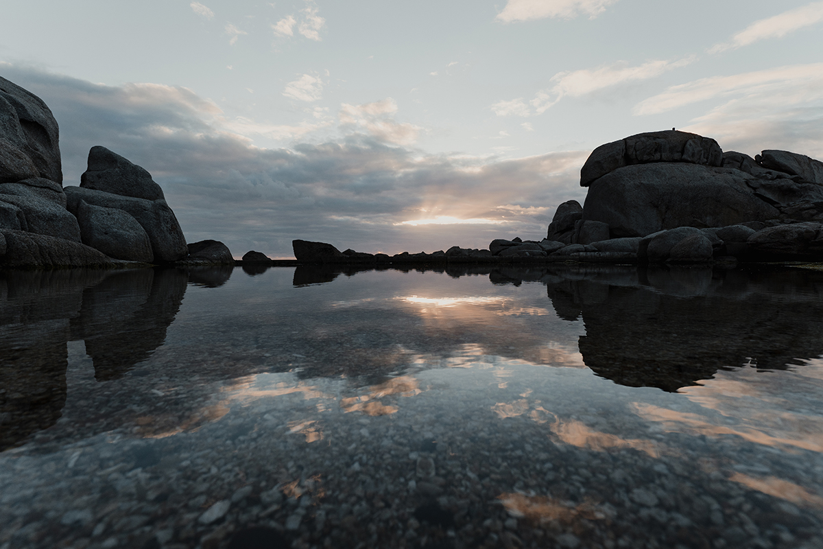 Subtle sunrise over rocky shoreline with still water reflecting clouds and sky, captured using the best settings for sunset and sunrise photos.