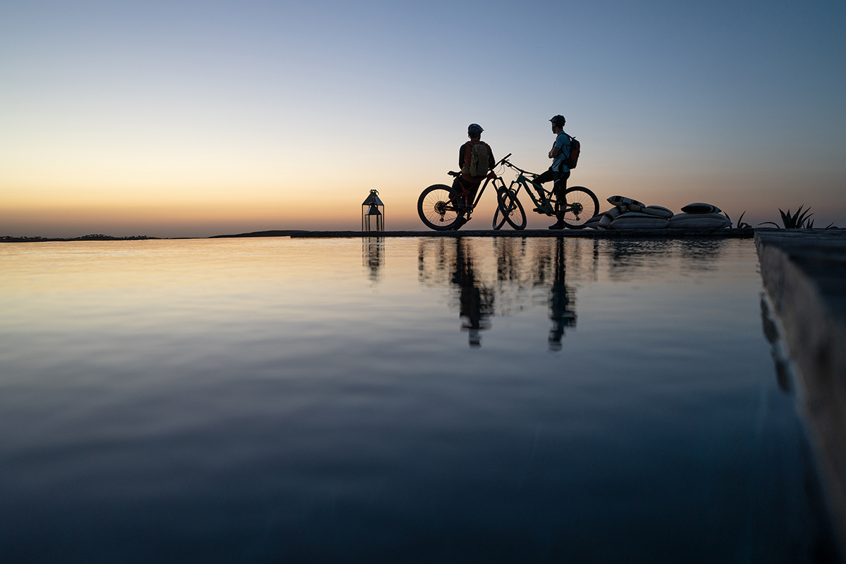 Silhouettes of two cyclists at sunrise near calm water with reflections and a lantern-lit platform in the background.