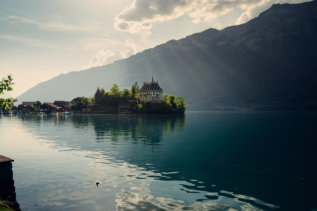Los rayos de sol se cuelan a través de las nubes sobre un lago sereno y una villa histórica, demostrando el dominio de las técnicas de iluminación de exteriores con luz natural suave y difusa. ©Ryad Guelmaoui