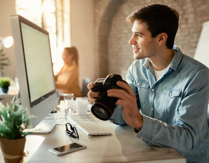 Um homem sentado em frente ao computador segurando uma câmera com uma lente