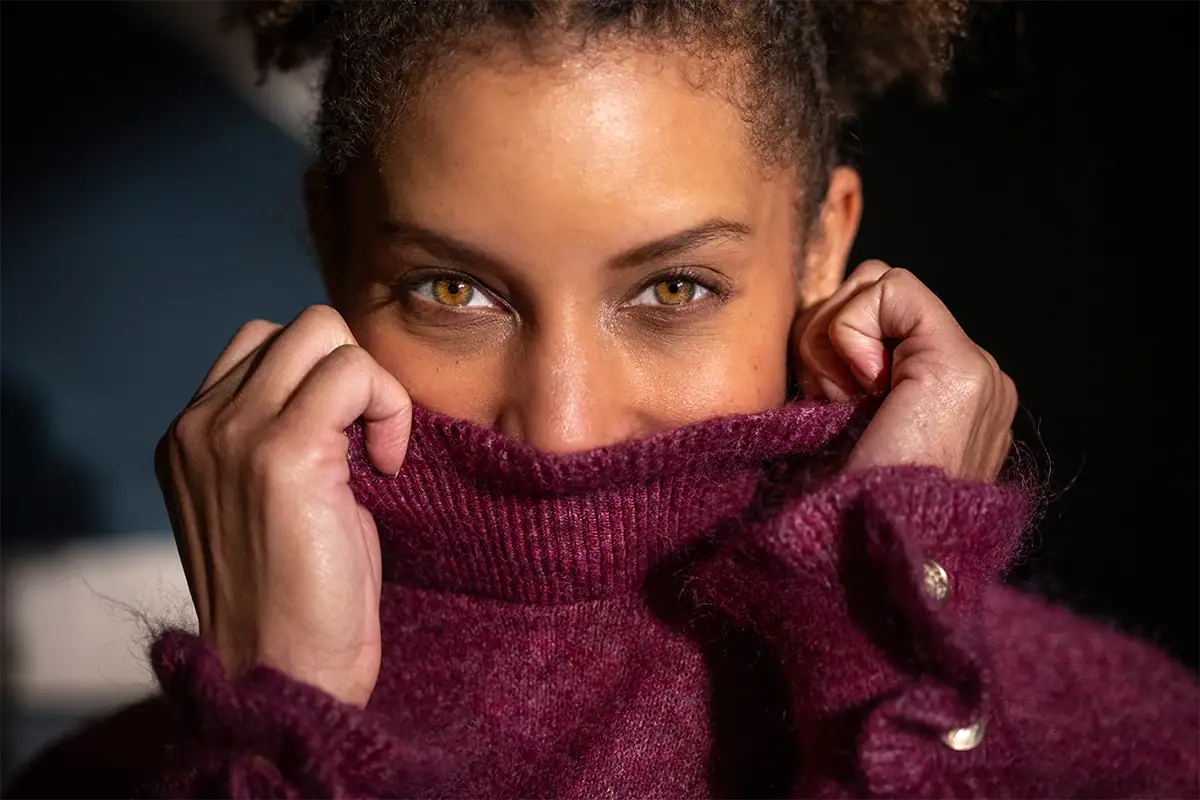 Close-up portrait of a woman partially covering her face with a burgundy sweater, showcasing sharp eye detail and smooth background blur captured with a Tamron 35-100mm F2.8 lens on a Sony A7R full-frame mirrorless camera.