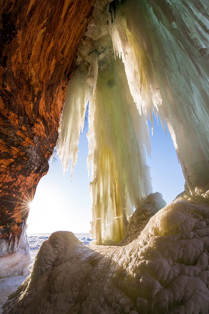 Pingentes de gelo iluminados pelo sol pendurados em uma caverna de rocha vermelha ao nascer do sol - demonstrando técnicas criativas de fotografia de inverno por meio de luz, textura e enquadramento.