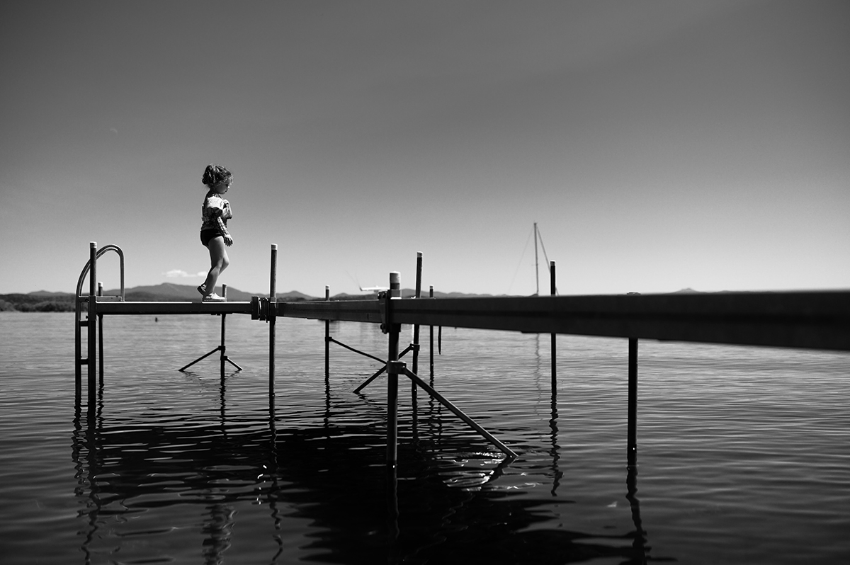 Foto em preto e branco de uma menina caminhando em uma doca sobre a água - mostrando como fotografar retratos de família com uma narrativa emocional e atemporal. ©Andre Costantini