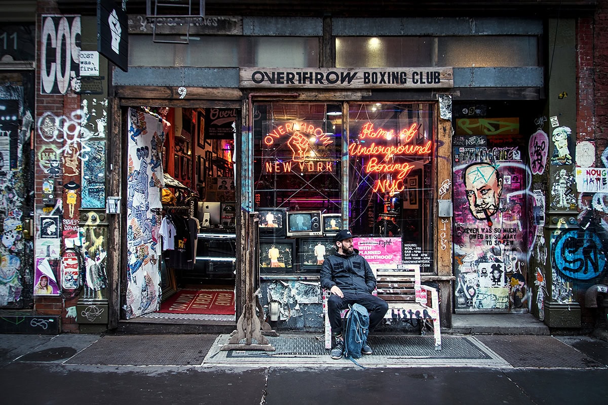 Un hombre sentado en el exterior del Overthrow Boxing Club de Nueva York, rodeado de vibrante arte callejero y letreros de neón: un ejemplo de aplicación de los consejos de la fotografía local para captar el auténtico carácter urbano.