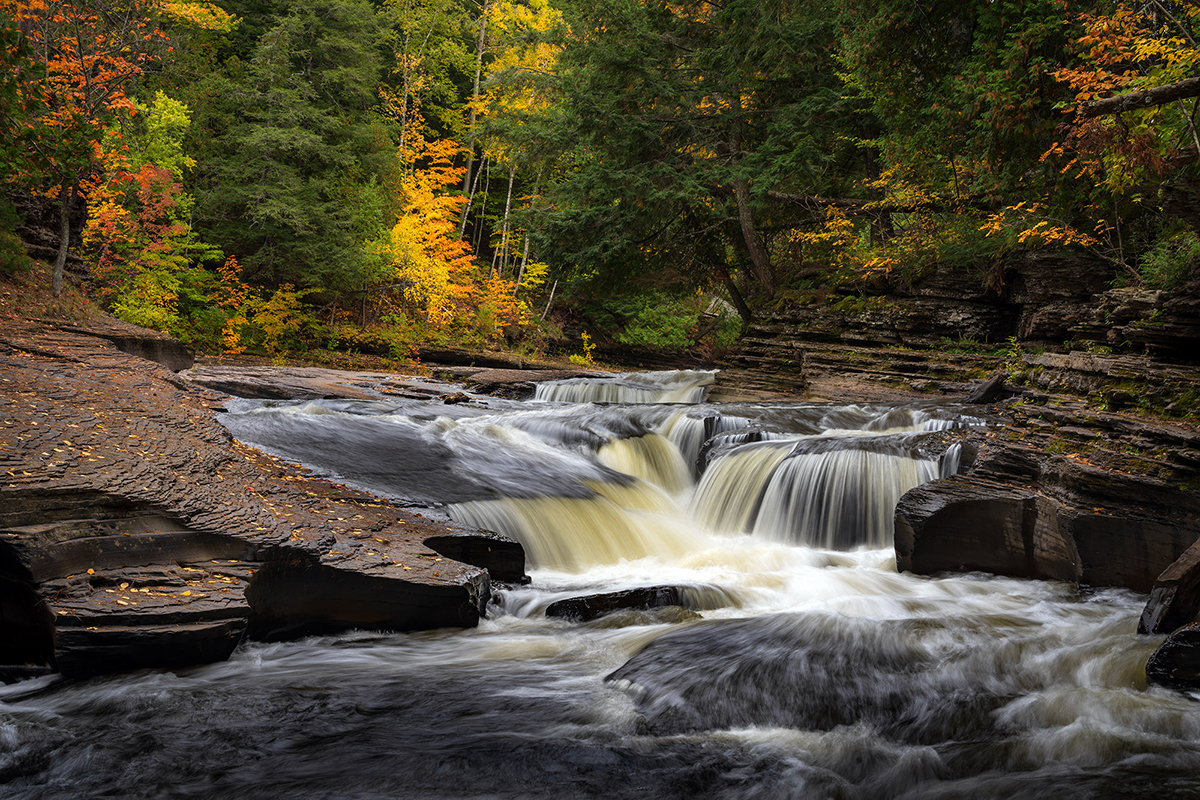 Dramatic landscape photo of a cascading waterfall in the Porcupine Mountains, surrounded by vibrant autumn foliage, captured with a Tamron zoom lens.