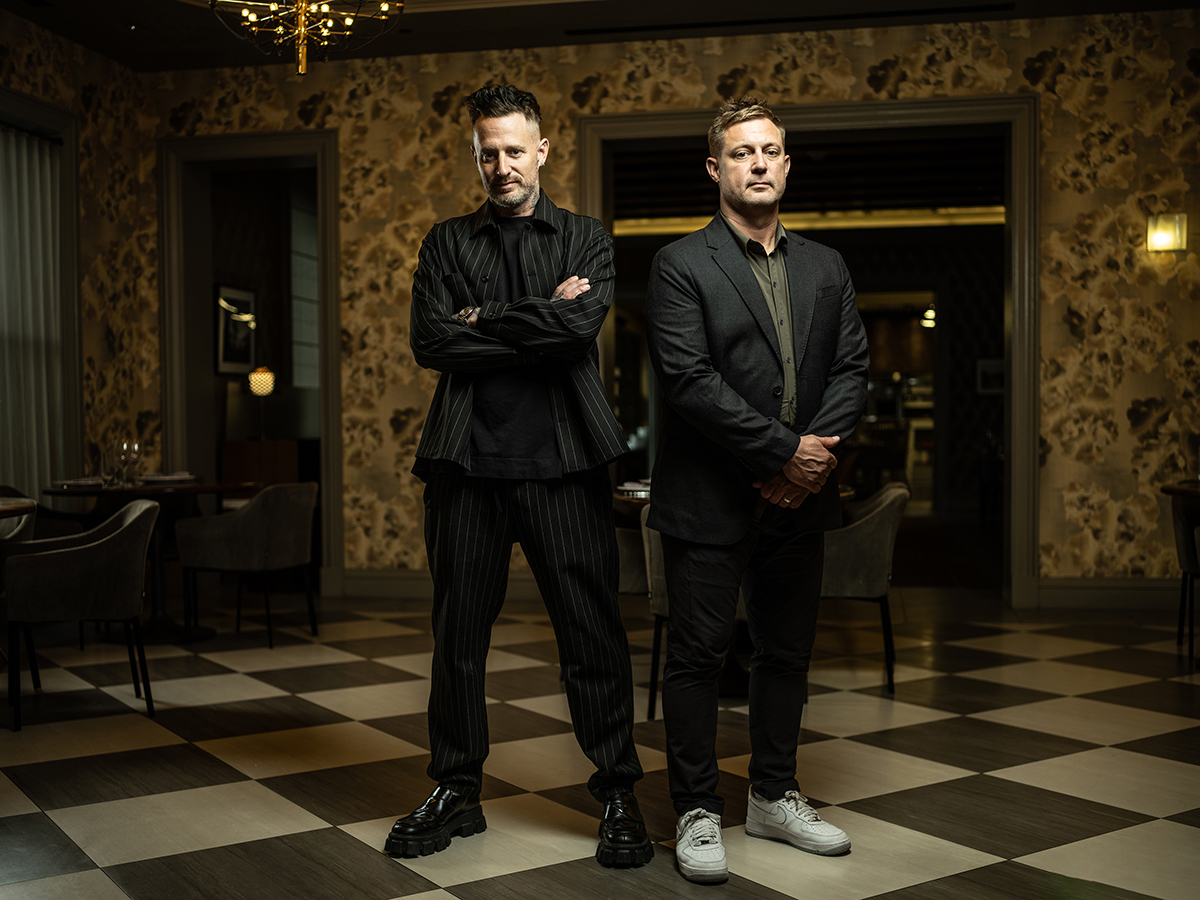 Dos hombres de pie con confianza en un interior sofisticado, demostrando una iluminación dramática en la fotografía de retrato con alto contraste y profundidad.