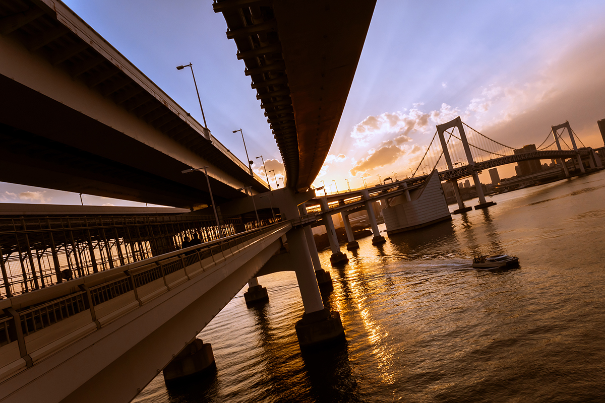Golden sunset light shining through bridge structures over a river, with a boat passing below and dramatic clouds in the sky.