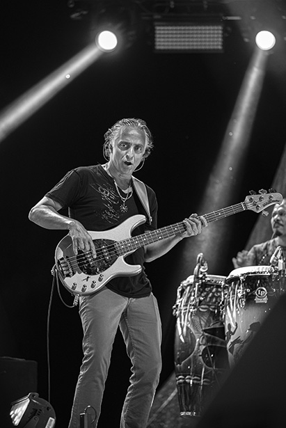 Bajista actuando en directo en el escenario con iluminación expresiva y percusionista de fondo.