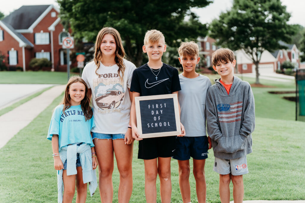 Groupe de cinq enfants souriants en âge d'aller à l'école se tenant ensemble à l'extérieur dans un quartier de banlieue, l'un d'entre eux tenant une pancarte sur laquelle on peut lire "Premier jour d'école" - un portrait amusant et candide de la rentrée des classes avec des amis.