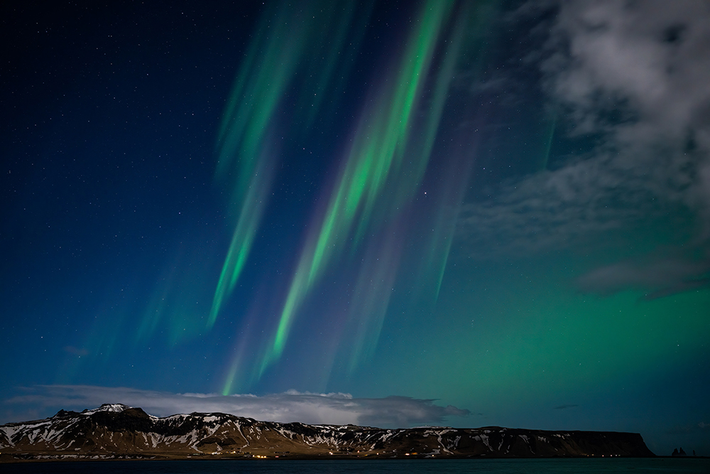 Vibrant green and purple aurora borealis streak across a starry night sky above a snow-dusted mountain range and coastal village in Iceland, with clouds partially illuminated by the aurora&rsquo;s glow.