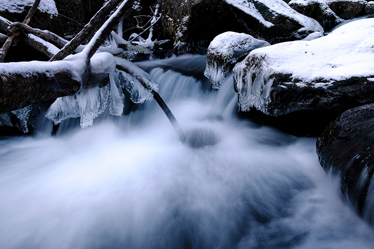 A winter scene showing a stream surrounded by icy trees and rocks, enhanced through long exposure photo editing techniques.
