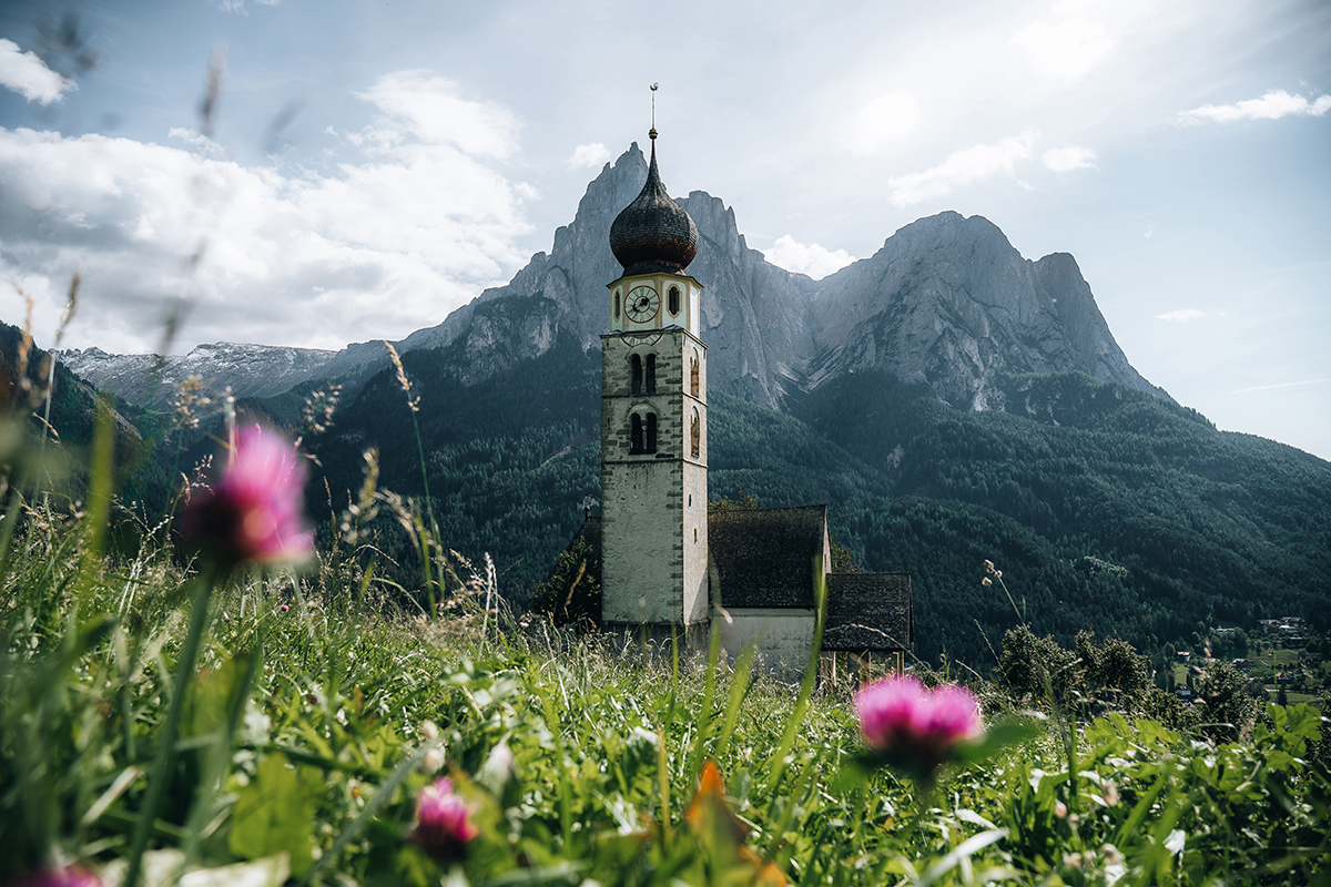 Igreja alpina e cenário de montanhas escarpadas emolduradas por flores silvestres, tirada com a lente Tamron 17-70 mm para paisagens.