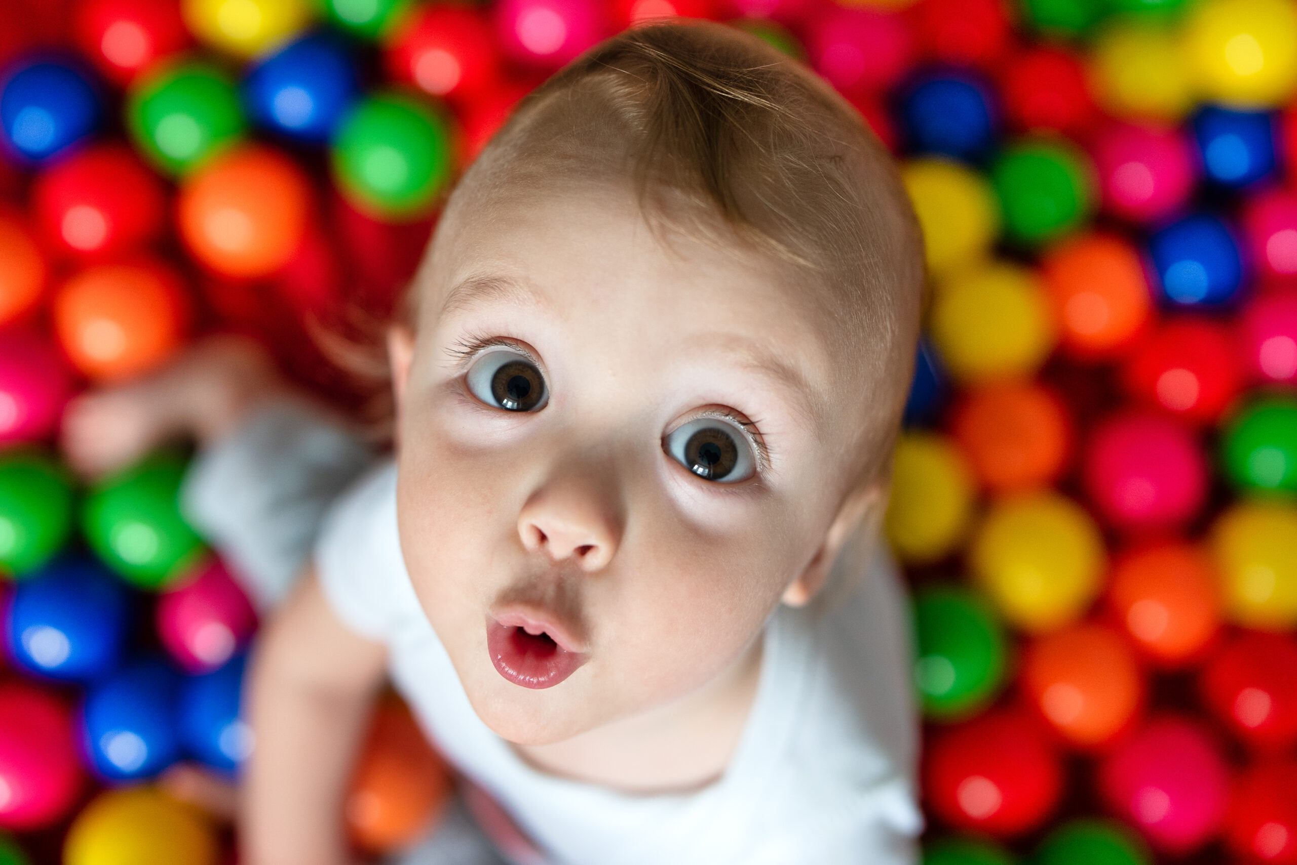 Criança de olhos arregalados em uma piscina de bolinhas coloridas olhando para cima com um rosto expressivo - capturando a emoção lúdica na fotografia de retratos de crianças.
