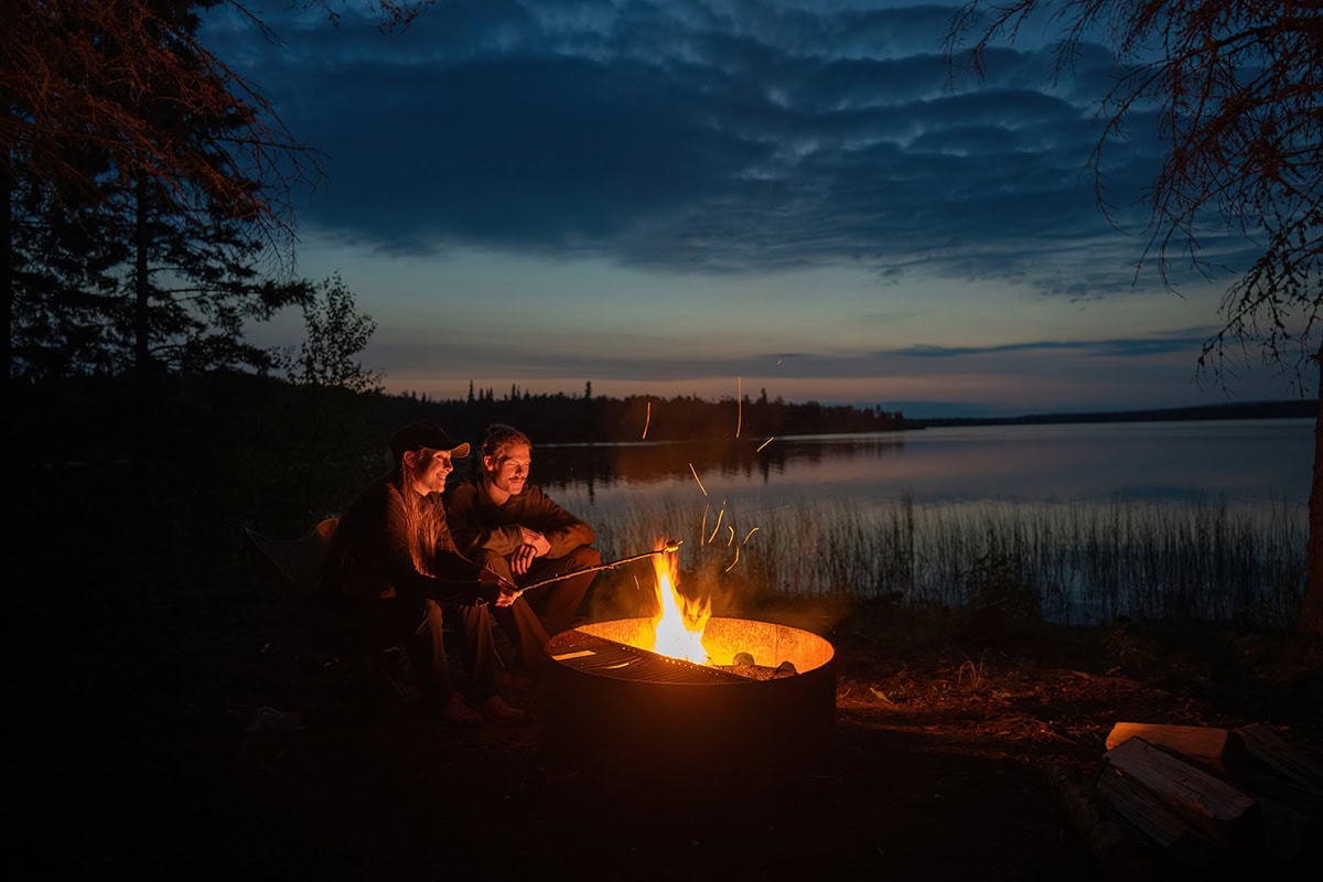 Dois campistas sentam-se perto de uma fogueira à beira de um lago ao anoitecer, demonstrando a iluminação ideal para fotos da hora dourada - perfeita para aplicar dicas de fotografia de acampamento.