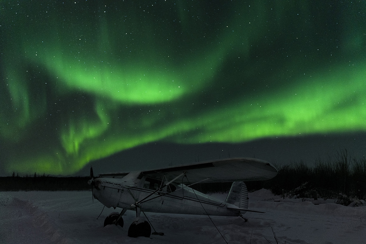 Aurora Boreal resplandeciente de verde en un cielo nocturno lleno de estrellas con un avión cubierto de nieve en primer plano, ilustrando una escena perfecta para la fotografía del cielo nocturno y consejos de fotografía de lluvia de meteoritos.