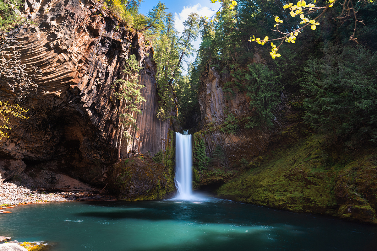 Fotografía de larga exposición de una cascada rodeada de bosque y formaciones rocosas, con agua suave y vibrantes colores diurnos.